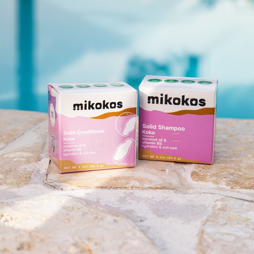 Cajas del shampoo sólido y el acondicionador sólido Koko de Mikokos sobre una piedra junto a una piscina