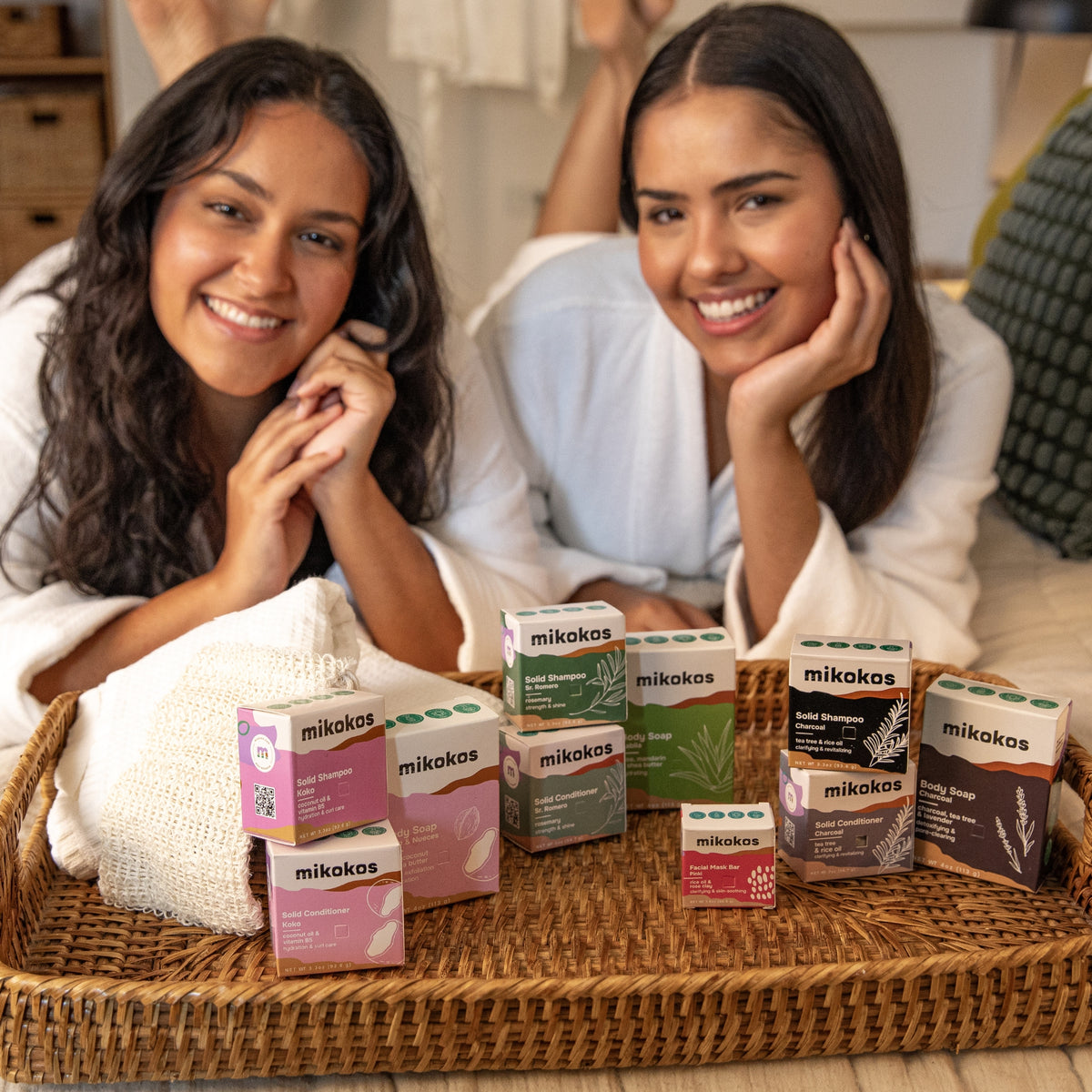 Bandeja con productos Mikokos (shampoo, acondicionador, jabón y mascarilla) junto a dos personas relajándose en batas, día de self-care en casa